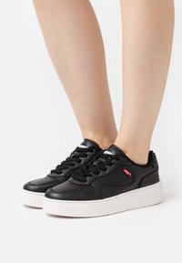 Baskets noires avec une tige en cuir lisse, détails perforés, bout rond, semelle en caoutchouc blanche contrastante et lacets noirs. Accents de logo rouge.