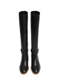 Botas altas de cuero negro con punteras redondeadas, detalle de correa cerca de los tobillos y suelas marrones, mostradas de frente sobre un fondo blanco.