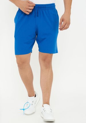 Mann trägt blaue Shorts mit elastischem Bund und weiße Sneakers, steht vor weißem Hintergrund und justiert den Bund mit der linken Hand.