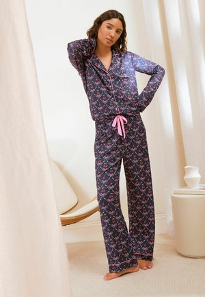 Kvinde står barfodet i mørkeblå mønstrede pyjamas med lyserødt bånd, poserer med den ene hånd på nakken og den anden på hoften indendørs.