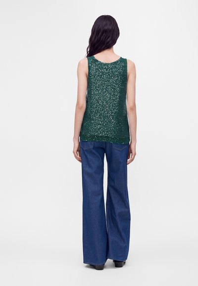 Femme aux longs cheveux foncés, portant un haut sans manches vert à sequins et un jean bleu à jambes larges, debout de dos sur fond blanc.