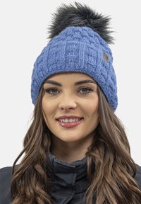 Căciulă tricotată albastră cu un model texturat și un pom-pom din blană sintetică neagră pe vârf. Include o mică etichetă logo din piele maro.