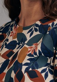 Blouse multicolore présentant un motif floral avec des feuilles et des baies. Le tissu a une texture lisse, avec des nuances de bleu, de vert et d'orange.
