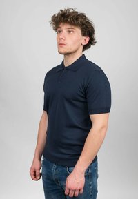 Námořnická modrá polo košile s krátkým rukávem, límečkem, žebrovanými manžetami a hladkou texturou. Model pózuje z boku v modrých džínách.