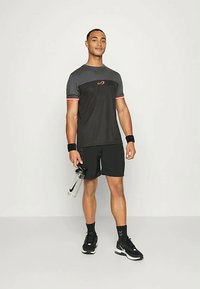 Fekete és szürke sportpóló rózsaszín kiegészítőkkel, fekete sorttal párosítva. A modell egy átlátszó vízpalackot tart, és fekete edzőcipőt visel.