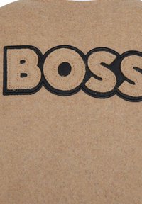 Tessuto in feltro beige con un grande logo ricamato in nero "BOSS". Il design è liscio con dettagli di cucitura visibili attorno alle lettere.