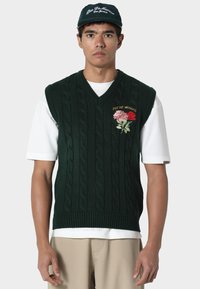 Vihreä palmikkoneuleliivi, jossa on V-aukko ja pinkin ja punaisen sävyinen kukkaommel. Päällä valkoinen t-paita ja resorireuna helman kohdalla.