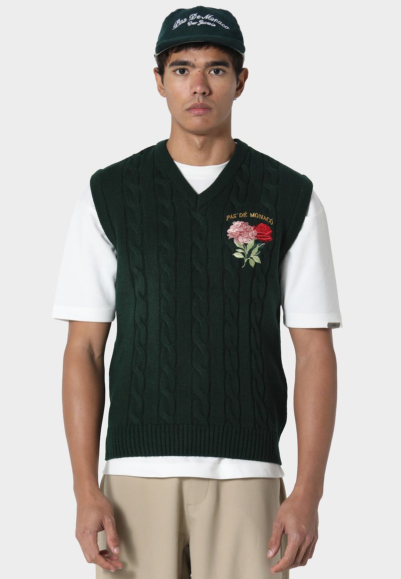 Vihreä palmikkoneuleliivi, jossa on V-aukko ja pinkin ja punaisen sävyinen kukkaommel. Päällä valkoinen t-paita ja resorireuna helman kohdalla.