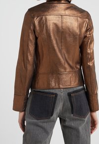 Bronzová kovová kožená bunda s přiléhavým střihem, viditelné švy a kombinována se šedými džíny s kontrastními tmavými kapsami.