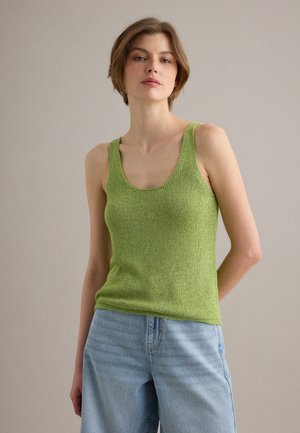 Femme portant un débardeur texturé vert clair sans manches et un jean bleu clair, debout devant un fond clair uni.