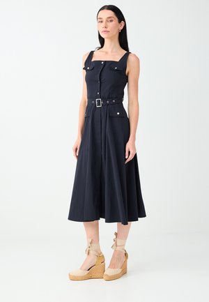 Femme portant une robe midi noire sans manches avec des boutons, une ceinture et des poches, associée à des espadrilles compensées beiges avec des liens à la cheville.