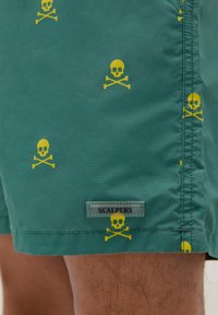 Shorts de bain verts avec motif de crâne et os croisés jaunes. Présente une étiquette de marque "SCALPERS" sur le côté. Texture lisse.