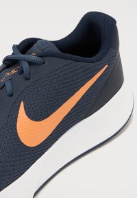 Marinblå idrettssko med strukturert mesh-overdel, oransje Nike swoosh-logo og hvit mellomsåle. Har grå detaljer og komfortable snører.