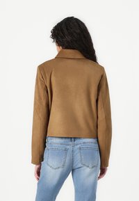 Hnědá umělá suede bunda s límečkem, zkráceným střihem a dlouhými rukávy, spárovaná se světle modrými džíny s zadními kapsami.