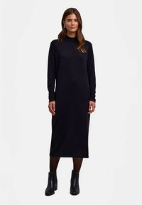 Zwarte jurk met lange mouwen, een hoge kraag, een nauwsluitende pasvorm en een ontwerp tot op de knie, gecombineerd met zwarte enkellaarzen en doorschijnende panty's.