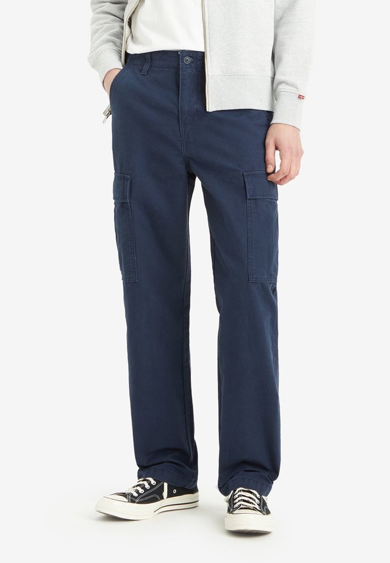 Marineblauwe cargobroek van katoen, met meerdere zijzakken, een losse pasvorm en een knoopsluiting. Draagt zwart sneakers.