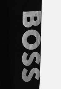 Juodos sportinės kelnės, ant kojos turinčios išryškintą sidabrinį "BOSS" logotipą, su glotnia tekstūra ir laisvalaikio stiliumi.