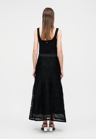 Femme aux longs cheveux bruns portant un haut noir sans manches, une jupe maxi en dentelle noire et des sandales noires à talons blocs blancs, debout de dos.