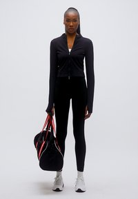 Μαύρο αθλητικό μπουφάν με φερμουάρ, μαύρο κολάν, λευκά αθλητικά παπούτσια. Κρατά μια μαύρη τσάντα duffle με κόκκινα διακοσμητικά. Λείο, εφαρμοστό ύφασμα.