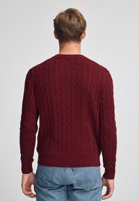 Burgundy kabelstickad tröja med rund halsringning, långa ärmar och ribbade ärmslut, med ett strukturerat mönster genom hela plagget.