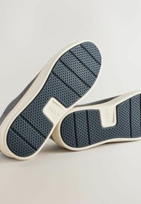 Suole di sneaker in navy con una superficie testurizzata e dettagli beige. Le suole presentano un motivo a rete per una migliore aderenza e un logo stampato in rilievo.
