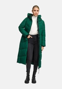 Woman wearing a long green hooded puffer coat over white sweater, black skinny jeans, and black ankle boots, standing with one hand in pocket.