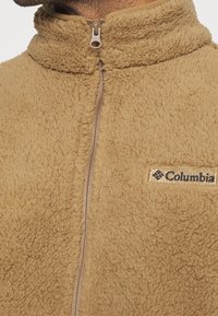 Beige fleecejacka med hög krage, hel dragkedja fram och Columbia-logotyp på vänster bröst. Texturerad, mjuk tyg.