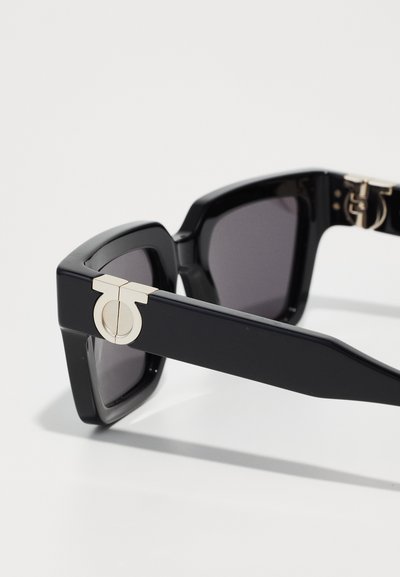 Black sunglasses with a thick, angular frame. Lenses are dark gray. Side features a gold circular accent and hardware. Smooth texture throughout.