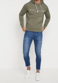 Uomo in felpa olive green di Tommy Hilfiger, jeans blu attillati arrotolati alle caviglie e sneaker bianche, in piedi con le mani vicino alle tasche.