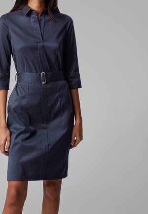 Femme portant une robe chemise bleu foncé ceinturée avec des manches trois-quarts sur un fond gris uni.