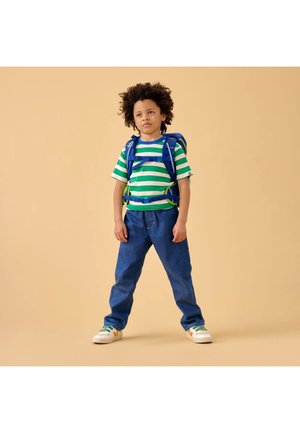 Child with curly hair wearing a green and white striped shirt, blue jeans, white sneakers, and a blue backpack standing against beige background.