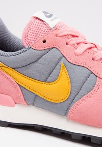 Nike tenisky s šedou síťovinou, růžovými semišovými akcenty a jasně žlutým logem swoosh. Obsahují růžové tkaničky a polstrovanou bílou podrážku.