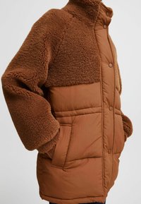 Brun jakke med en fluffy øverste sektion og en glat, quilted nederste sektion. Den har trykknapper og sidelommer.