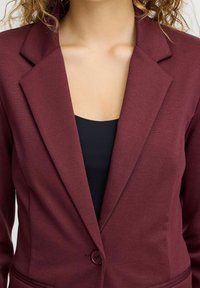 Burgundi bleiseri, jossa on viistetty kaulus, yksittäinen napin suljenta ja kaksi etutaskua, tehty teksturoidusta kankaasta. Musta toppi alla.