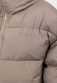 Giacca puffer beige con un design trapuntato, caratterizzata da un sottile logo ricamato e una cerniera a contrasto. Texture liscia e opaca.