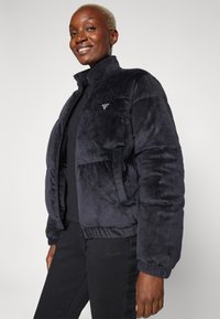 Giacca a vento nera con collo alto, realizzata in tessuto morbido. Presenta un piccolo logo sul lato sinistro del petto e polsini elasticizzati.