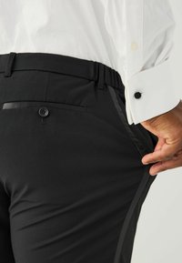 Melni bikses ar strukturētu apdari, ar sānu svītrām un aizmugures kabatu ar pogas aizdari. Valkātas ar baltu garām piedurknēm kreklu.