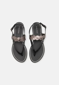 Svarta platta sandaler med en flätad metallrem, smala justerbara vristremmar och vit kant längs fotsidans kanter.