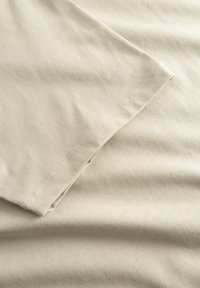 Beige bomuldsstof med en glat overflade, prydet med en foldet kant og subtile sømme. Ren finish uden synlige mønstre eller detaljer.