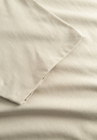Tissu en coton beige avec une texture lisse, présentant un bord plié et des coutures subtiles. Finition propre sans motifs ni accents visibles.