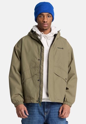 Junger Mann mit blauer Mütze, beiger Kapuzenjacke, hellgrauem Kapuzenpullover und blauen Jeans, vor einem einfarbigen Hintergrund stehend.