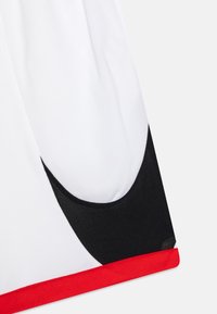 Vita idrottsshorts i vitt med svarta och röda accenter, featuring en mesh-textur, böjda designelement och ett kontrasterande rött midjeband.