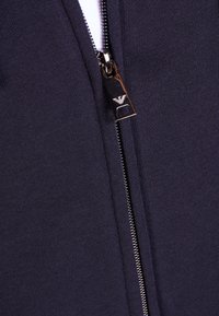 Marinblå zip-up hoodie med en metallisk dragkedja och logotypdragare. Slät stickad textur med en nära passform runt halslinningen.