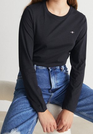 Vrouw draagt een zwart shirt met lange mouwen en een klein logo op de borst en een blauwe high-waist spijkerbroek, zittend met haar handen rustend tussen haar knieën.