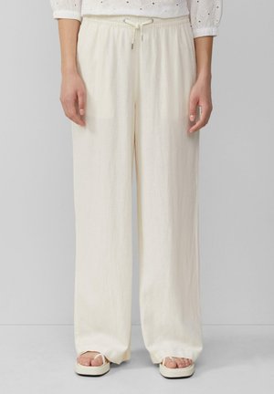 Volné kalhoty s vysokým pasem v krémové barvě, široké nohavice s tkanicí, nošené s bílými platformovými sandály a bílou halenou s vyšívanými dírkami.