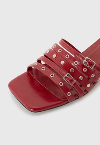 Sandalia de deslizamiento sintética roja con múltiples tiras horizontales, tachuelas de metal plateado y un diseño de punta cuadrada. Presenta un detalle de hebilla ajustable.