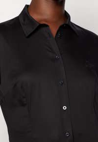 Schwarzes Hemd mit Knopfleiste aus glattem Stoff, mit klassischem Kragen, fünf Knöpfen und dezent gesticktem Logo auf der Brust.