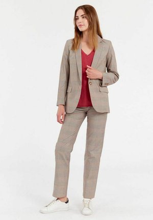 Costume à carreaux gris avec une veste ajustée et un pantalon assorti, associé à un haut rouge en V et des baskets blanches. Tissu lisse avec un design structuré.