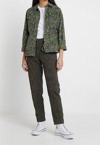 Veste verte à motif camouflage sur une chemise blanche, associée à un pantalon olive et des baskets blanches. La veste a une coupe décontractée et des poches à boutons.