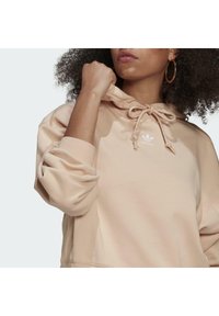 Ljust beige hoodie med dragsko i huvan, oversize ärmar och en liten vit Adidas-logotyp på bröstet. Mjuk tygtextur.